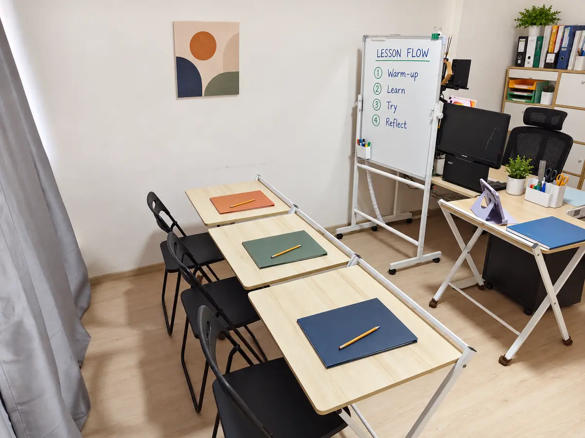 A home-classroom teaching zone with three learner desks, a lesson-flow whiteboard and organised teacher resources.